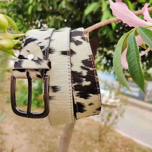 Black and white cow print belt with stitched edges and a dark matte pin buckle hanging from a tree branch with green leaves and pink flowers