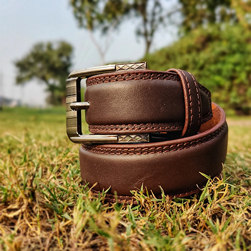 Men's brown leather belt coiled on dry green grass with double red-brown stitched edges and an antique pin buckle with grooved frame and crosshatch detail