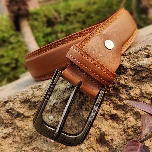 Men's tan leather belt with double yellow-orange stitched edges, engraved gunmetal pin buckle and gold-tone rivet resting on a rock outdoors